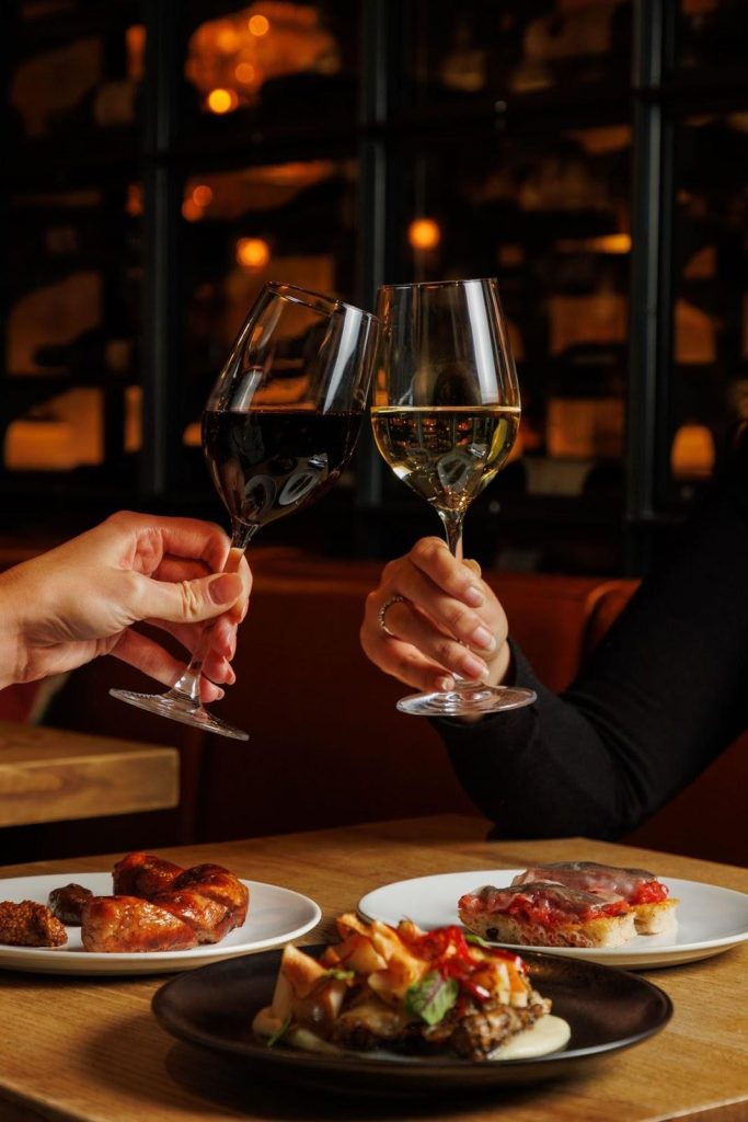 Two glasses of red wine being clinked over some dishes at a wine bar