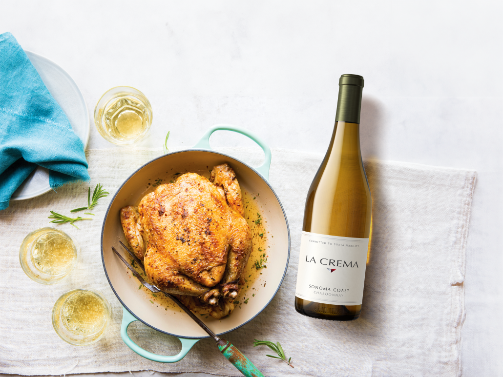A whole cooked chicken served next to a bottle and glasses of white wine