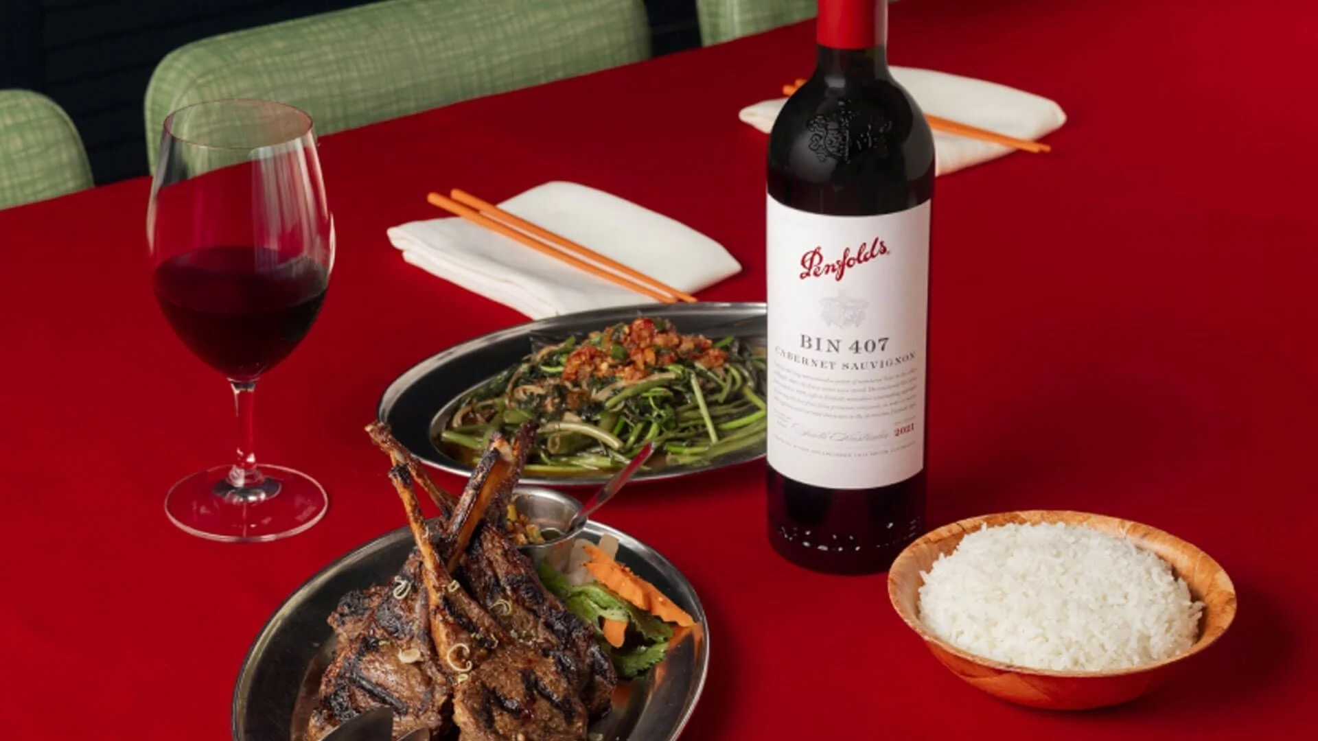Cooked meat, greens and rice dishes next to a glass and bottle of red wine